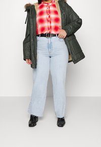 Kobieta ubrana w jasnoniebieskie dżinsy, czarne buty, czerwoną koszulę w kratę z guzikami oraz otwartą ciemnozieloną kurtkę z beżowym podszyciem.