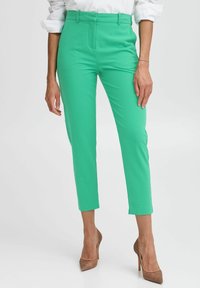 Pantalons ajustés verts lumineux avec une coupe slim, munis de passants de ceinture, de poches latérales et d'une texture lisse. Portés avec des talons hauts nude.