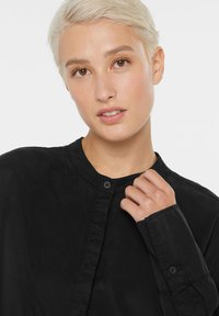 Schwarze Button-Down-Bluse aus strukturiertem Stoff, mit Rundkragen und langen Ärmeln mit geknöpften Manschetten. Nahaufnahme, die das Detail zeigt.