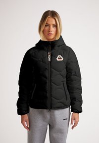 Giacca piumino nera con design trapuntato, chiusura con zip, cappuccio e toppa con logo. Abbinata a pantaloni della tuta grigi, con cuciture visibili e tasche.