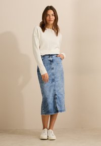 Leichter, cremiger Pullover kombiniert mit einem hellblauen Jeansrock in mittellanger Länge, der einen Schlitz vorne und Taschen aufweist. Abgerundet mit weißen Sneakers.