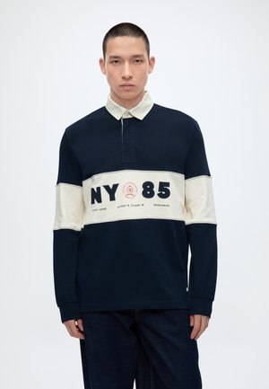Junger Mann mit kurzen Haaren, der ein marineblaues und cremefarbenes langärmeliges Polohemd mit der Aufschrift "NY 85" und dunkle Hosen trägt, vor weißem Hintergrund steht.