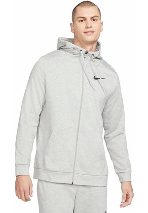 Homme portant un sweat à capuche gris clair zippé et un pantalon de survêtement assorti avec le logo Nike, debout devant un fond blanc.