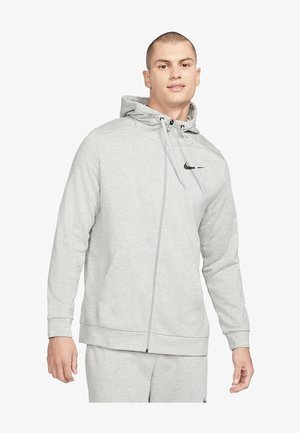 Hombre con sudadera con capucha gris claro con cremallera y pantalones deportivos a juego con el logo de Nike, de pie frente a un fondo blanco.