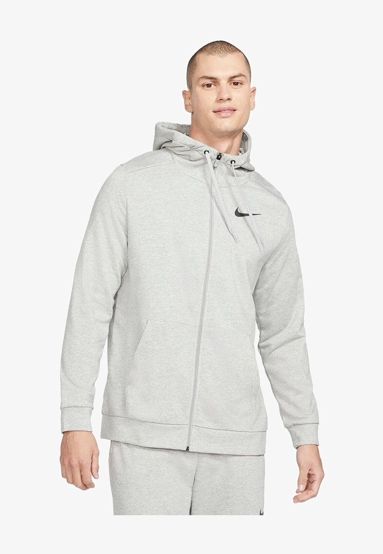 Homme portant un sweat à capuche gris clair zippé et un pantalon de survêtement assorti avec le logo Nike, debout devant un fond blanc.