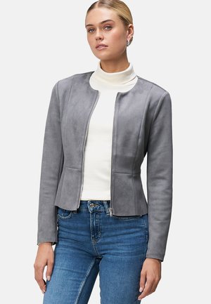 Veste en suédine grise avec un design ajusté, col rond et fermeture éclair, associée à un col roulé blanc et un jean bleu.