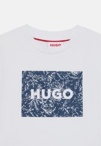 Λευκό t-shirt με κοντά μανίκια που διαθέτει ένα ναυτικό ορθογώνιο γραφικό με υφή και τη λέξη "HUGO" με έντονα λευκά γράμματα. Κόκκινη λεπτομέρεια στο λαιμό.