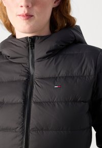 Black puffer jacket with a zip-up front and a hood. Features horizontal quilting and a small logo in red, white, and blue on the chest.
