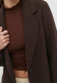 Donkerbruin wollen blend jas met een notch-kraag, gestructureerde schouders en een enkele knoopsluiting, gelaagd over een bijpassend bruin top.