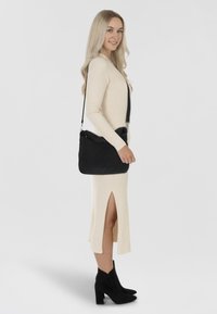 Crni teksturirani crossbody torba sa zatvaračem na vrhu, nošena na ramenu. Model nosi krem pleteni haljinu sa bočnim prorezom i crne gležnjače.