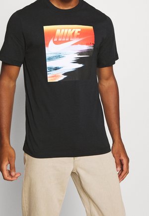 Persona che indossa una maglietta nera Nike con una grafica colorata di un tramonto sulla spiaggia e pantaloni beige.