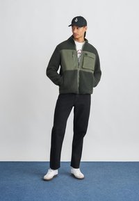Green quilted fleece jacket with a zip-up front, two-tone design, chest pocket, paired with black pants and white sneakers.