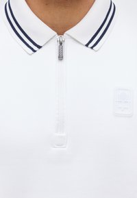 Λευκό πόλο με φερμουάρ, γιακάς με ναυτικές ρίγες και φερμουάρ με το λογότυπο Lacoste, με διακριτικό ανάγλυφο έμβλημα ρακέτας του τένις στο στήθος.