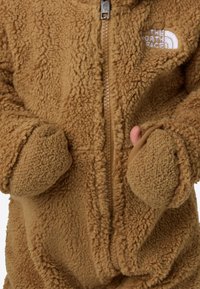 Béžová flísová bunda s textúrovaným povrchom, celým zipsom a dvoma vreckami. Na ľavej hornej strane je biely vyšívaný logo.