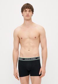 Melnas bokseros ar pieguļošu dizainu, kas izceļas ar kontrastējošu jostu, uz kuras ir balti un zaļi uzraksti "JACK & JONES".