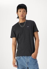 Mørkegrå t-skjorte med korte ermer og rund hals, med en liten logo på brystet, iført lyseblå baggy jeans.