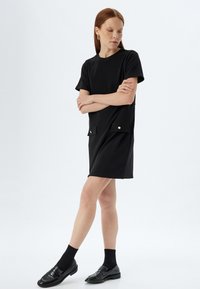 Zwart kort shirtjurkje van zacht materiaal, met twee voorzakken met gouden knopen. Gemaakt in combinatie met zwarte sokken en gepolijste schoenen.
