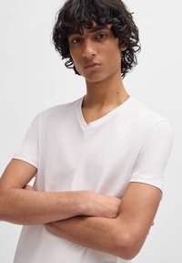 White V-neck T-shirt in soft cotton, short sleeves, fitted design. Model's arms crossed, curly hair, neutral background.