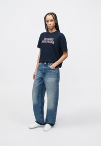 Μπλε ναυτικού χρώματος μπλουζάκι με το λογότυπο "TOMMY HILFIGER" σε λευκό και κόκκινο. Φωτεινά γαλάζια τζιν ψηλόμεσα. Λευκά αθλητικά παπούτσια. Λείο ύφασμα και άνετη εφαρμογή.