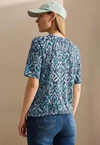 T-shirt blu a maniche corte con un motivo verde e blu navy, realizzata in tessuto morbido. Indossata con jeans in denim scuro e un cappellino verde chiaro.