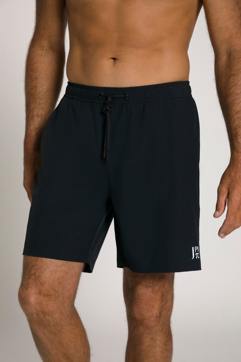 Uomo che indossa pantaloncini neri con coulisse e un piccolo logo bianco sulla parte inferiore della gamba sinistra, in piedi davanti a uno sfondo semplice.