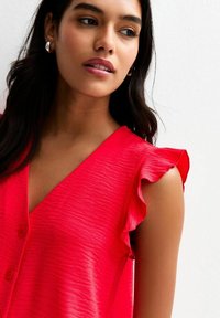 Haut sans manches texturé rouge avec un col en V et des accents de volants sur les épaules. Présente trois boutons sur le devant.