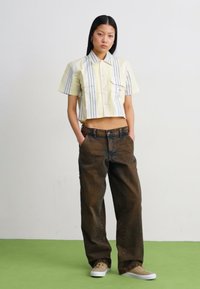 Camicia gialla a righe con maniche corte abbinata a jeans marroni larghi e scarpe beige. Presenta tasche e un design dalla vestibilità rilassata.
