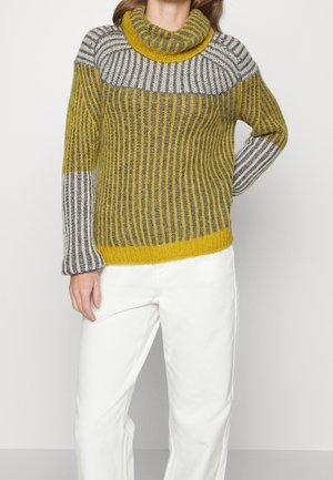 Žena stojící s jednou rukou v bok, oblečená v pruhovaném žlutém, šedém a bílém pleteném roláku a bílých kalhotách.