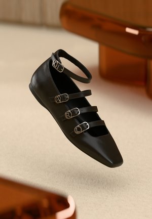 Chaussure plate en cuir noir avec un bout pointu, dotée de trois sangles ajustables et de boucles métalliques. Texture lisse et design minimaliste.