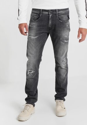 Homme portant un jean noir usé avec des bottes blanches à lacets et une chemise blanche à manches longues, debout devant un fond uni.
