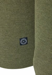 Khaki zielony sweter, strukturyzowany splot, żebrowany dół, z okrągłą granatową etykietą z białym tekstem, umieszczoną w dolnym lewym rogu.