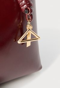 Geantă de mână din piele burgundă cu suprafață netedă, având o bretea împletită și un charm-logo triunghiular auriu pentru un plus de detaliu.