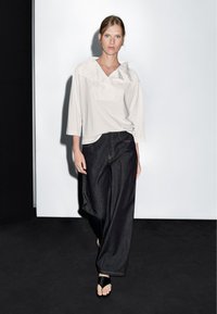 Světle šedý, volně přiléhající top s širokým výstřihem a předním skladem; spárovaný s tmavě modrými, širokými džínovými kalhotami. Jednoduché černé sandály dokončují vzhled.