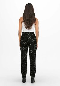 Woman with long brown hair wearing a white sleeveless top and black polka dot pants, standing facing away on a white background.