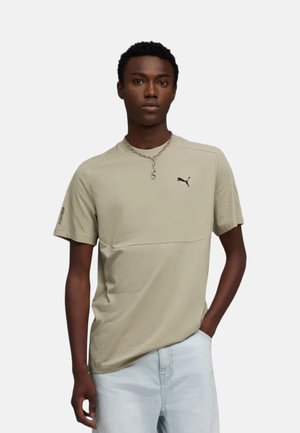 Joven con camiseta beige de Puma y vaqueros azul claro, de pie con una mano en el bolsillo frente a un fondo liso.