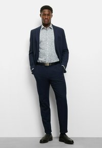 Marineblauer Anzug mit schlanker Passform und strukturiertem Stoff. Hellblaues Hemd mit floralem Muster darunter. Schwarzer Gürtel und polierte Schuhe runden das Outfit ab.