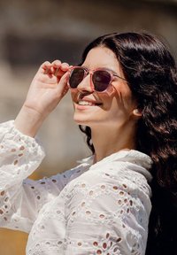 Růžové kulaté sluneční brýle s průhledným rámem; bílá vyšívaná blůza s nabíranými rukávy. Osoba se usmívá, zatímco si upravuje brýle.