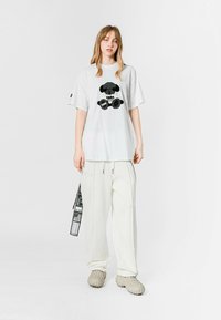 Camiseta blanca de gran tamaño con un gráfico de un oso de peluche negro, combinada con pantalones blancos holgados y zapatos texturizados en un beige claro.