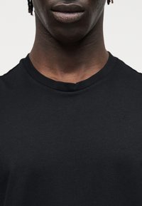 Κοντινό πλάνο του κάτω μέρους προσώπου και του λαιμού ενός ατόμου που φοράει απλό μαύρο μπλουζάκι με στρογγυλή λαιμόκοψη, με φόντο έναν απλό φωτεινό τοίχο.