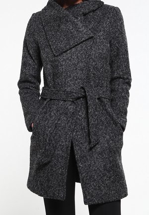 Mørkegrå tekstureret uldfrakke med bred krave, bælte i taljen og hænder i lommerne, båret over sorte bukser.