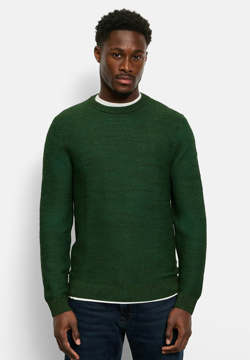 Grüner Strickpullover mit strukturiertem Muster, Rundhalsausschnitt und gerippten Bündchen. Über einem weißen Shirt getragen, kombiniert mit dunklen Jeans.