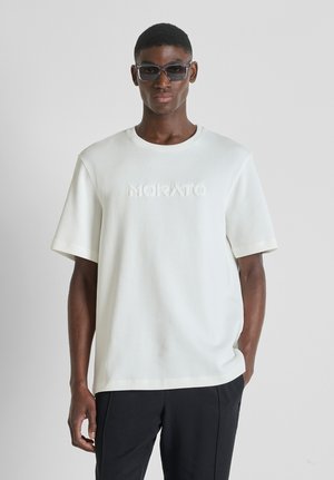 Muškarac u prevelikoj bijeloj majici s reljefnim logotipom "MORATO", crnim hlačama i tamnim pravokutnim sunčanim naočalama, stoji uz svijetlu pozadinu.