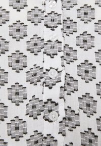 Tela blanca con un patrón geométrico negro, que presenta pequeñas formas similares a flores y botones de plástico transparente a lo largo del centro.