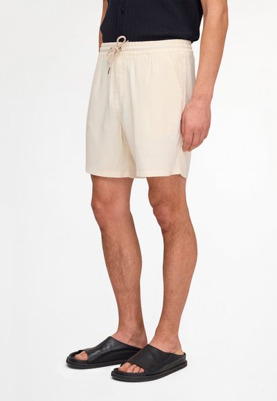 Male model wearing beige drawstring shorts and black open-toe slide sandals, standing against a white background.