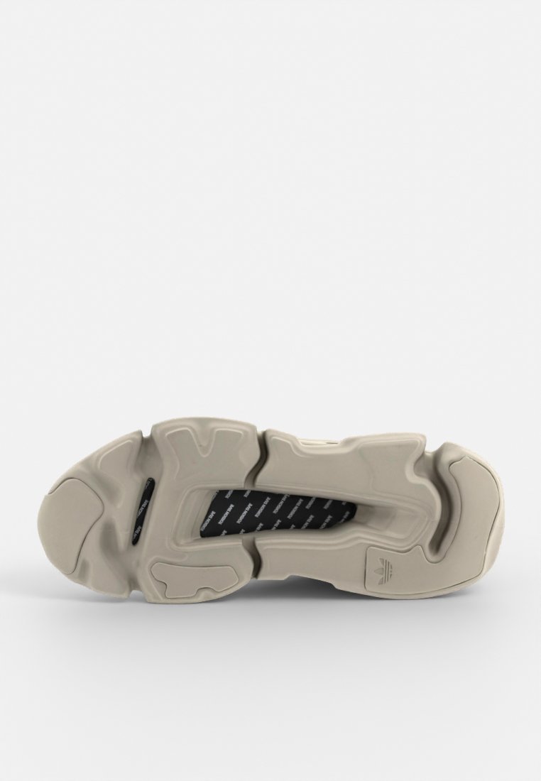 Suela de zapatilla deportiva en color crema con superficie de goma texturada, que presenta un diseño ondulado único y un forro negro interno con una marca blanca.
