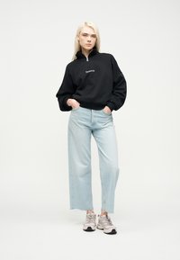 Νέα γυναίκα στέκεται με τα χέρια στις τσέπες, φοράει μαύρο φούτερ με μισό φερμουάρ, ανοιχτόχρωμα φαρδιά τζιν και μπεζ αθλητικά παπούτσια σε λευκό φόντο.