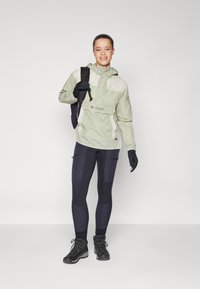 Lys grøn og kremet jakke med hætte, parret med sorte tights og handsker. Model bærer sorte vandrestøvler og har en rygsæk på.