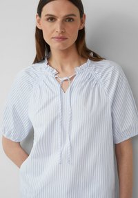 Blouse rayée bleu clair et blanche avec un col froncé, un détail en forme de trou de serrure et des manches courtes bouffantes. Fabriquée en tissu lisse.