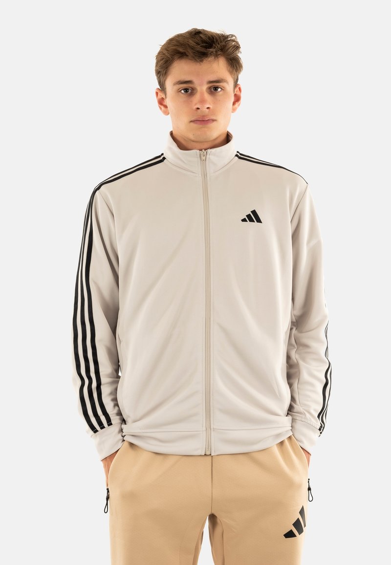 Chaqueta de chándal con cremallera en gris claro y detalles en negro. Presenta tres franjas negras a lo largo de las mangas y un logotipo bordado en el pecho.