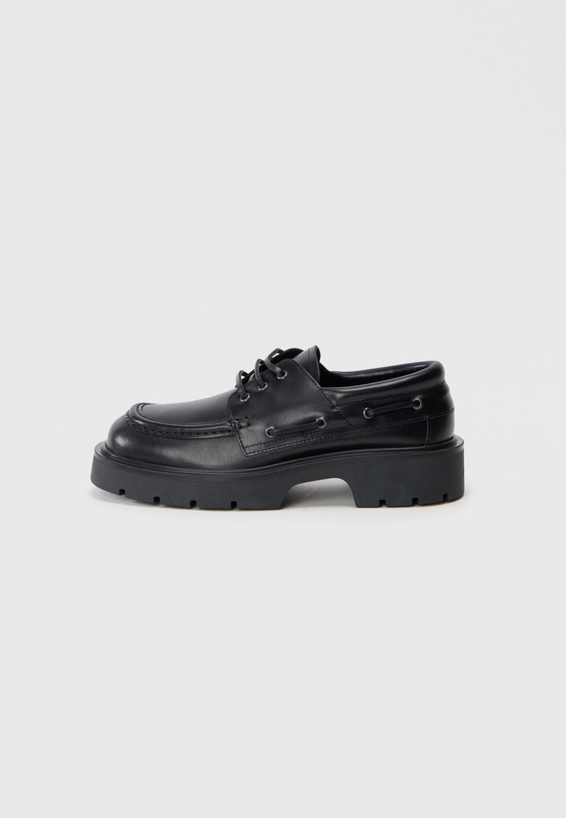 Schwarze Lederschuhe mit dicker Sohle im Boot-Stil, geflochtenem Schnürsenkel und Mokassin-Stich auf einem schlichten hellen Hintergrund.